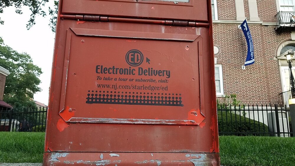 An empty red newspaper box carries a notice directing readers to the publication’s website.